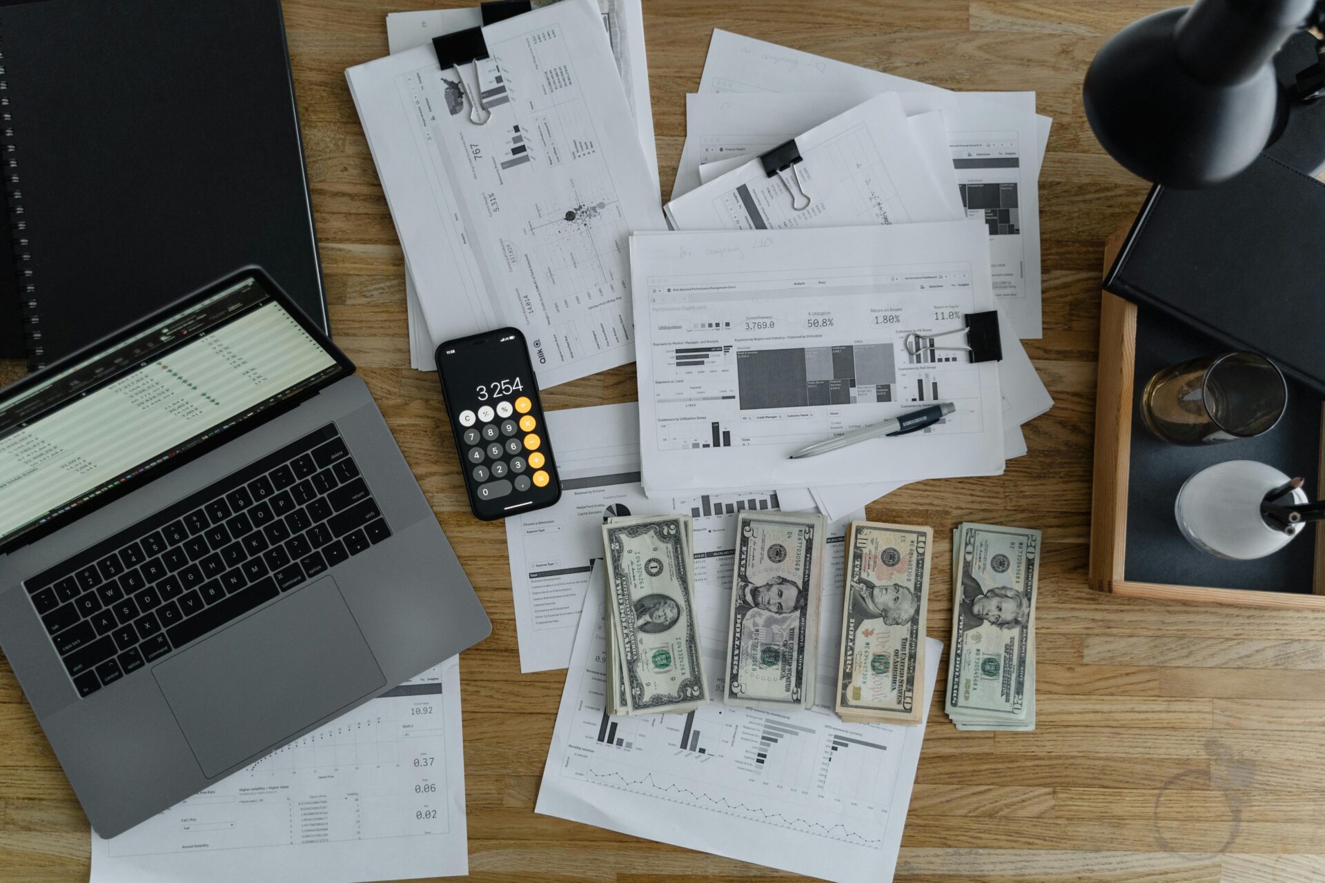 A desk filled with financial reports, a laptop, calculator, and stacks of cash, representing small business cash flow management during summer slowdown.